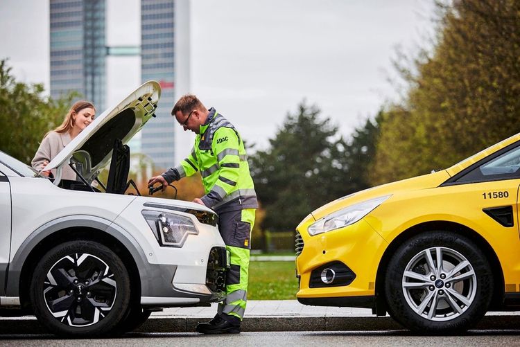 Ein ADAC-Mitarbeiter in gelb-grüner Warnkleidung hilft bei einer Fahrzeugpanne. Er verbindet ein gelbes ADAC-Fahrzeug mit einem weißen Auto mittels Starthilfekabeln. Im Hintergrund sind ein moderner Bürokomplex und Bäume zu sehen.