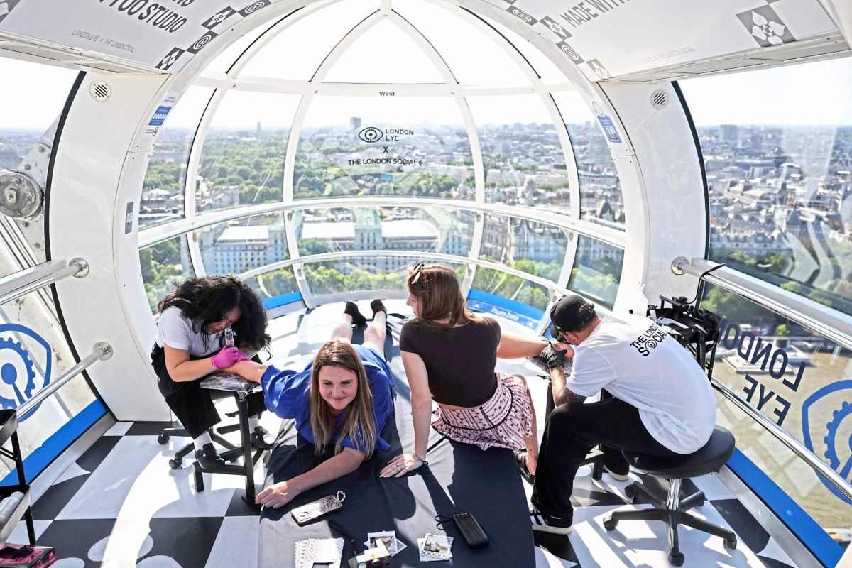 In einer Kabine des London Eye tätowieren die Künstler*innen Chariz Joy Calendey (CJ) und Lucas Scardua zwei Personen, Meg Gianforte (links) und Abi Wright (rechts), während sie auf dem Boden und auf einem Hocker sitzen. Die Kabine bietet eine weite Aussicht auf London. Die Szene ist Teil einer Touristenpromotion.