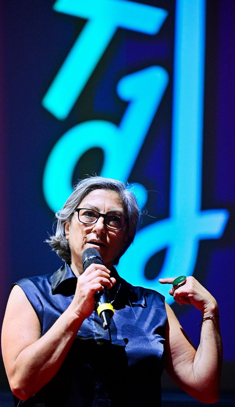 Eine Frau mit grauen Haaren und Brille spricht in ein Mikrofon und gestikuliert. Im Hintergrund ist ein blau-grüner Schriftzug mit den Buchstaben 