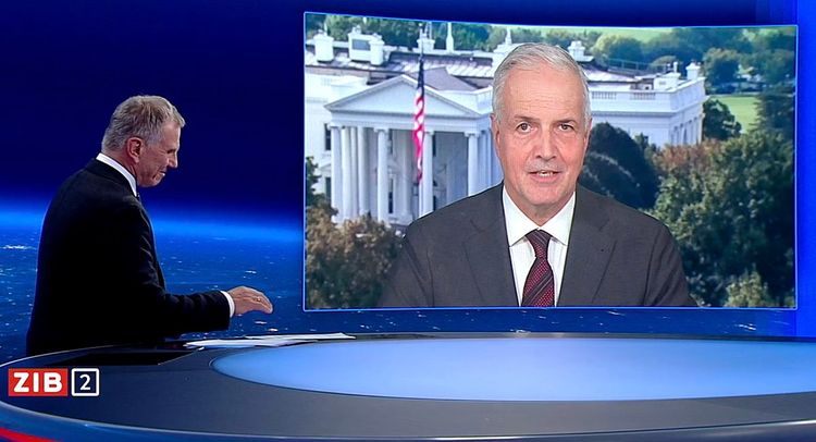 Mann in dunklem Anzug mit grauen Haaren spricht mit einem Mann mit dunkelgrauem Sakko, Krawatte, hellgrauen Haaren auf einem Display, im Hintergrund ist das Weiße Haus eingeblendet.