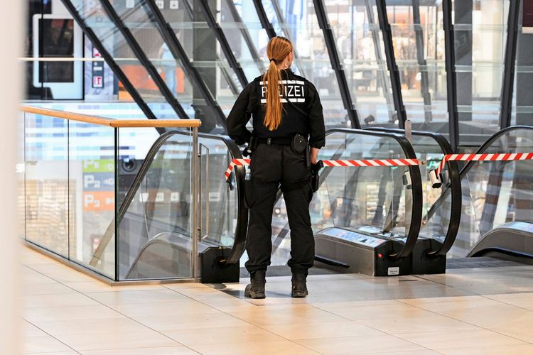 Eine Polizistin in schwarzer Uniform steht in einem Einkaufszentrum vor einer mit Absperrband gesicherten Rolltreppe. Im Hintergrund sind Glasfronten und Schilder mit Parkhinweisen erkennbar.