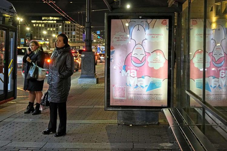 Frauen stehen neben einer Bushaltestelle in der nächtlichen Großstadt St. Petersburg, die Anzeigentafel zeigt eine Information für Schwangere. 