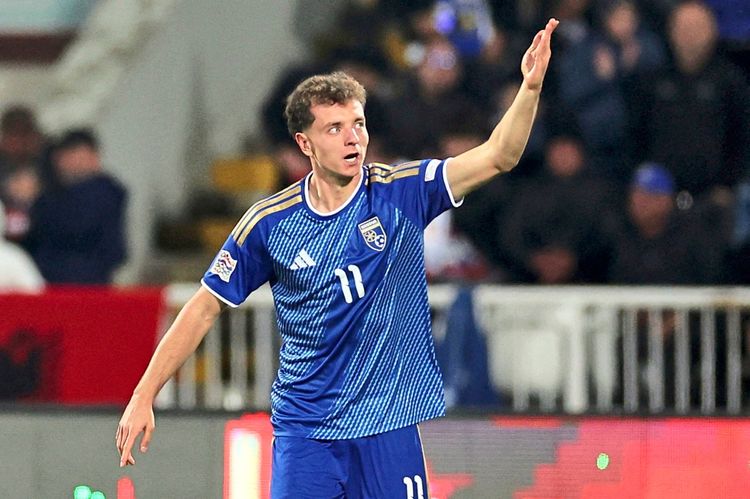 Ein Fußballspieler im blauen Trikot der kosovarischen Nationalmannschaft jubelt und hebt eine Hand. Der Hintergrund zeigt unscharfe Zuschauer im Stadion.