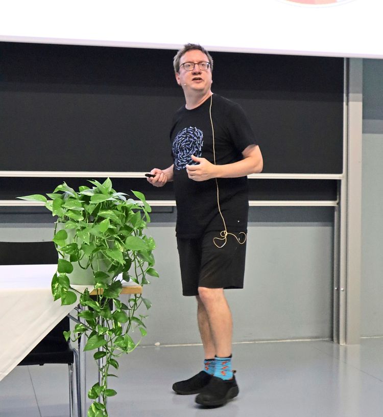 Ein Mann hält einen Vortrag während der ECMTB26-Konferenz. Er trägt ein schwarzes T-Shirt, schwarze Shorts und blaue Socken mit Mustern. Er steht vor einer Tafel und einem Projektorbild mit einem Kreisdiagramm. Links ist eine Pflanze auf einem mit weißer Tischdecke bedeckten Tisch zu sehen.