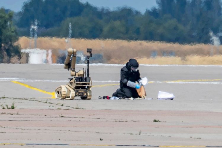 Eine Sicherheitskraft in Schutzausrüstung kniet auf einem Flugfeld und hantiert mit Gegenständen. Neben ihm ein unbemanntes Hilfsfahrzeug, das offenbar zum Entschärfen von Sprengsätzen eingesetzt werden kann.