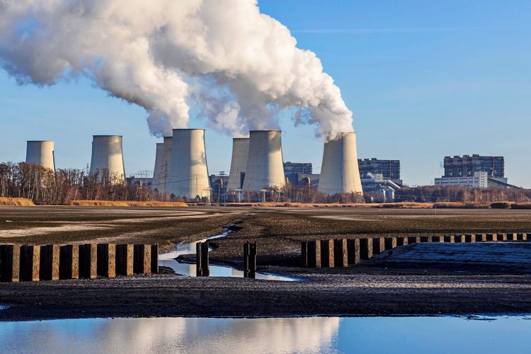 Braunkohlekraftwerk Jänschwalde in Brandenburg, Deutschland, mit mehreren dampfenden Kühltürmen, gesehen über eine Teichlandschaft mit Wasserflächen und Holzpfosten im Vordergrund. Klarer Himmel im Hintergrund.