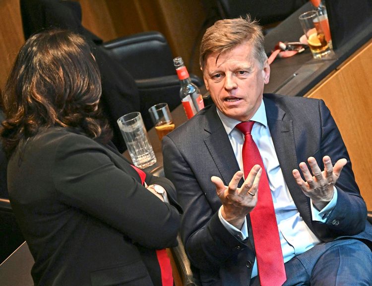 Ein Mann in einem Anzug mit roter Krawatte sitzt in einem Gespräch mit einer Frau mit dunklen Haaren in einem schwarzen Blazer. Im Hintergrund sind Gläser und Flaschen auf einem Tisch zu sehen.