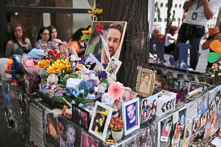 Eine Gedenkstätte mit Fotos, Blumen, Kerzen und Erinnerungsstücken ist um einen Baum arrangiert. Im Hintergrund sind Menschen versammelt, einige sitzen, andere stehen. Die Szene zeigt eine emotionale Gedenkveranstaltung.