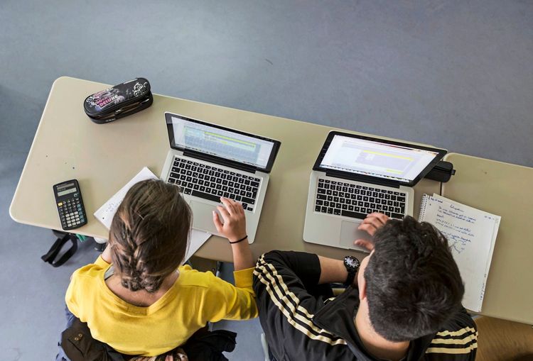 Zwei Schüler sitzen an einem Tisch und arbeiten jeweils an Laptops. Auf dem Tisch liegen ein Taschenrechner, Notizblätter und ein Federmäppchen. Der Raum hat einen schlichten grauen Boden. Szene aus einer Schulklasse.