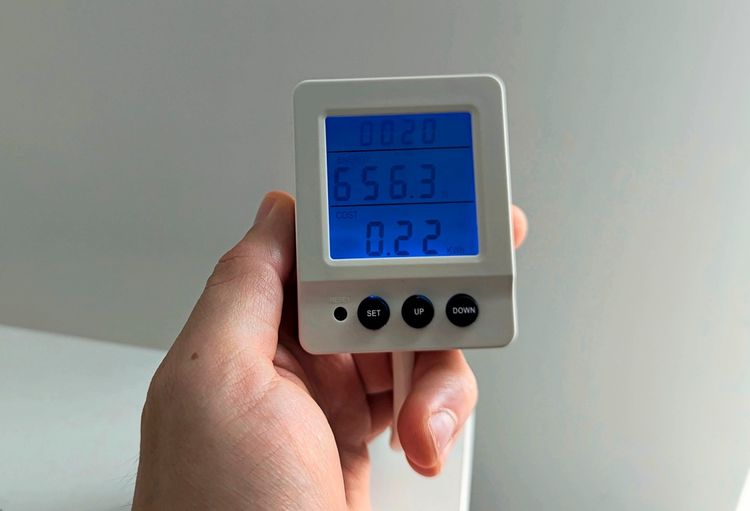Eine Hand hält ein weißes Energiekostenmessgerät mit blau beleuchtetem Display. Auf dem Display sind Werte wie Energieverbrauch (656,3 W) und Kosten (0,22 kWh) angezeigt. Darunter befinden sich vier schwarze Tasten: 