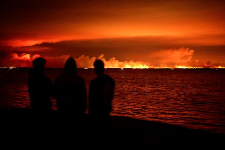 Drei silhouettierte Personen beobachten Waldbrände in der Ferne über einer Gewässerbucht bei Nacht. Der Himmel ist von intensiven orangefarbenen Flammen und Rauchwolken erleuchtet.