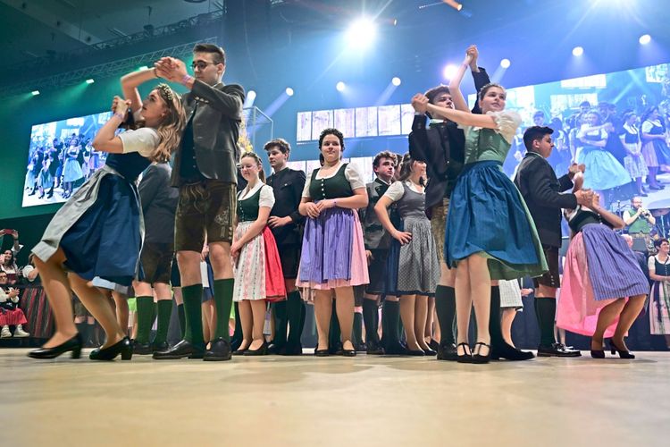 Eine Gruppe von Menschen in traditioneller Tracht tanzt auf einem Ball. Die Frauen tragen Dirndl, die Männer Lederhosen. Im Hintergrund sind Bühnenbeleuchtung, Bildschirme und weitere Teilnehmer zu sehen. Festliche Stimmung herrscht.