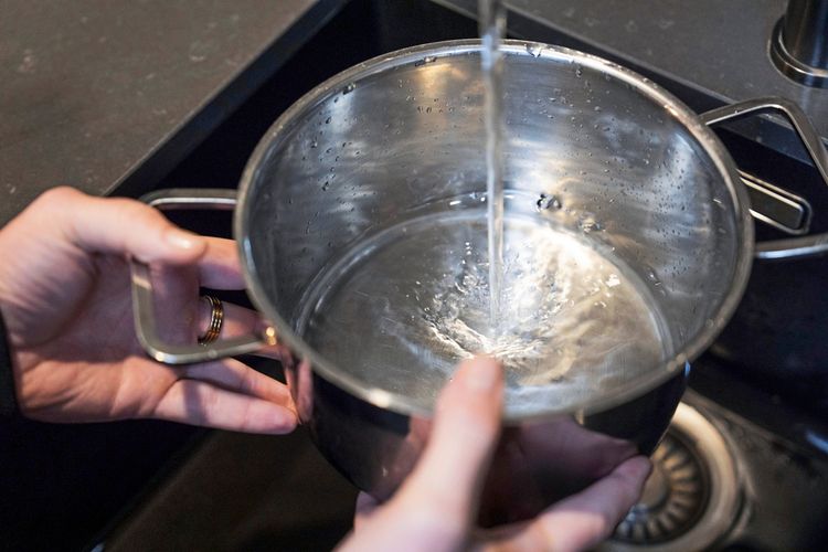 Hände halten einen Topf unter einen Wasserhahn, während Leitungswasser hineingefüllt wird.