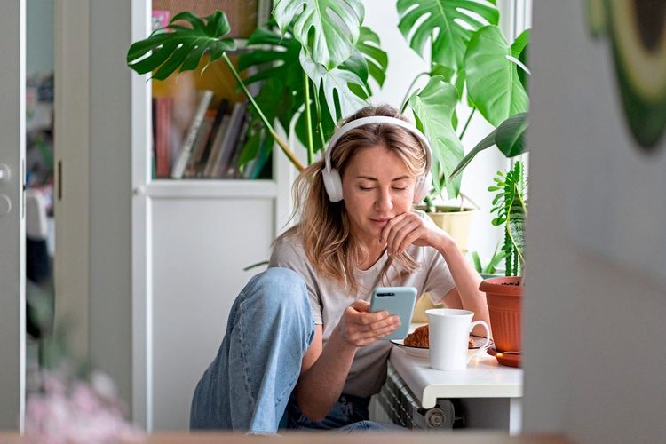 Frau sitzt mit einer Tasse Kaffee und einem Croissant auf einer Fensterbank, schaut auf ihr Smartphone und hört mit Kopfhörern Musik