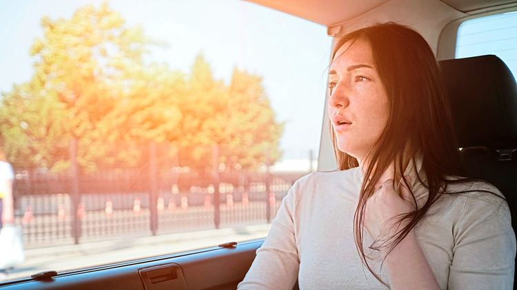 Eine Frau sitzt im Auto und sieht blass aus, die Hand in einer gestressten Geste an den Hals gelegt, sie zerrt an ihrem Halsausschnitt