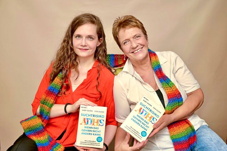 Zwei Frauen sitzen nebeneinander vor einem beigen Hintergrund und lächeln in die Kamera. Beide tragen einen bunten, regenbogenfarbenen Schal. Sie halten zusammen ein Buch mit dem Titel „Suchtrisiko ADHS – Wenn das Gehirn nicht anders kann“ in den Händen.