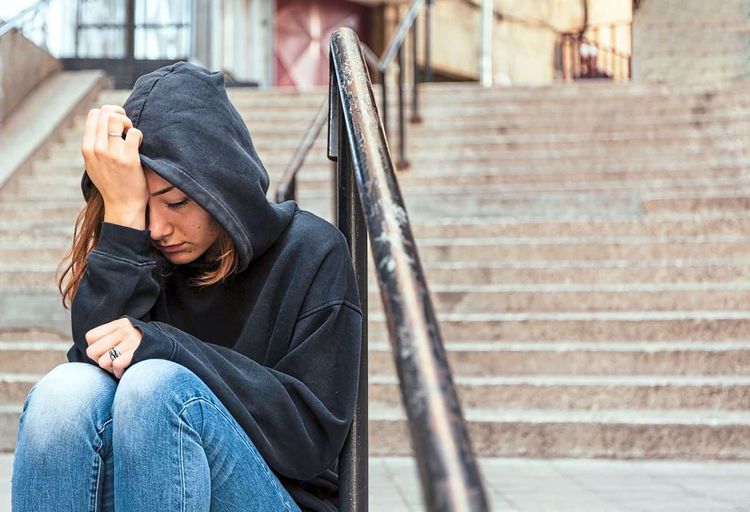 Mädchen im Teenager-Alter sitzt in schwarzem Kapuzenpullover und Jeans draußen auf einer Stiege und stützt den Kopf verzweifelt in die Hände