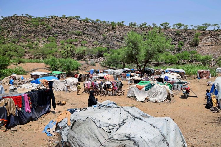 Ein provisorisches Lager von Binnenvertriebenen (IDPs) im al-Kadah-Gebiet, Taiz-Gouvernement, Jemen. Zelte aus Tüchern und Planen sind auf einer sandigen Fläche errichtet. Menschen bewegen sich im Lager, und Kleidung hängt zum Trocknen an den provisorischen Unterständen. Im Hintergrund sind hügeliges Gelände und Bäume sichtbar.
