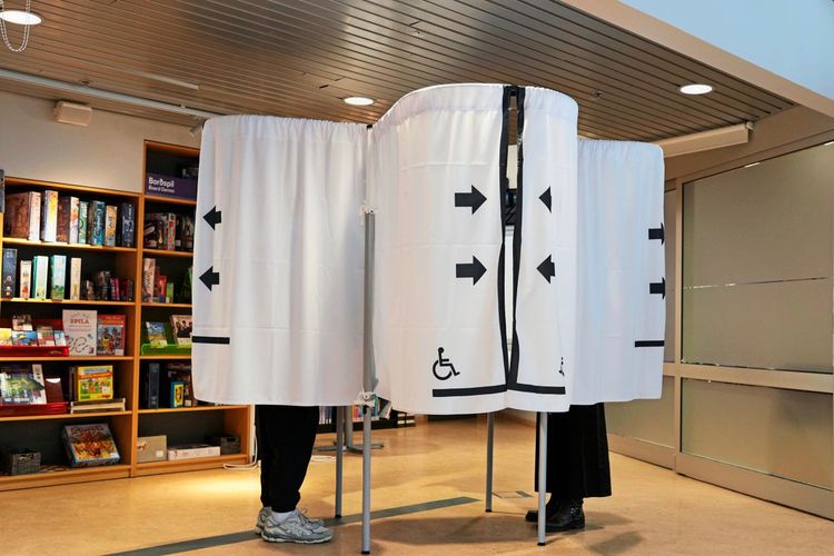 Ein Wahlraum in Reykjavik, Island, mit einer Wahlkabine, die von einem weißen Vorhang mit schwarzen Pfeilen und einem Rollstuhlsymbol verdeckt ist. Zwei Personen stehen hinter der Kabine, ihre Beine sind sichtbar. Im Hintergrund sind Regale mit Brettspielen zu sehen.
