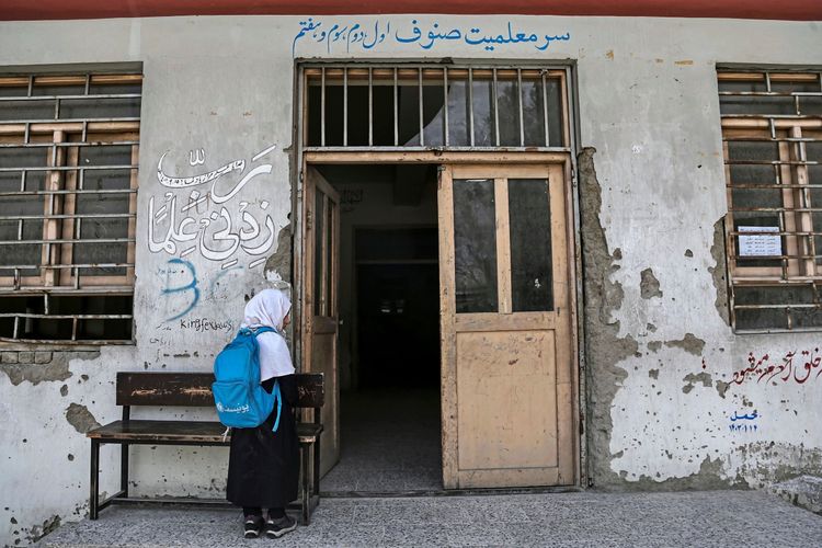 Ein afghanisches Schulmädchen mit einem blauen Rucksack steht vor dem Eingang einer Schule in Kabul, Afghanistan, am 22. März 2025. Die Wände des Gebäudes sind abgenutzt, mit Graffiti und Texten in einer regionalen Schrift versehen. Das Foto thematisiert die Bildungssituation von Mädchen in Afghanistan.