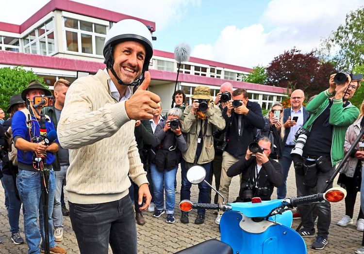 Ein Mann mit weißem Helm und beigem Pullover zeigt lächelnd einen Daumen nach oben, während hinter ihm mehrere Fotografen und Kameraleute stehen. Im Vordergrund ist ein blauer Motorroller zu sehen. Die Szene spielt sich vor einem Gebäude ab.