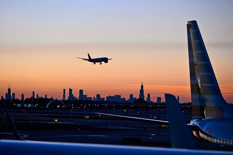 Ein Flugzeug landet bei Sonnenuntergang auf einem Flughafen mit der Skyline von Chicago im Hintergrund. Der Himmel ist in warmen Farben getönt, und Teile anderer Flugzeuge sind im Vordergrund sichtbar.