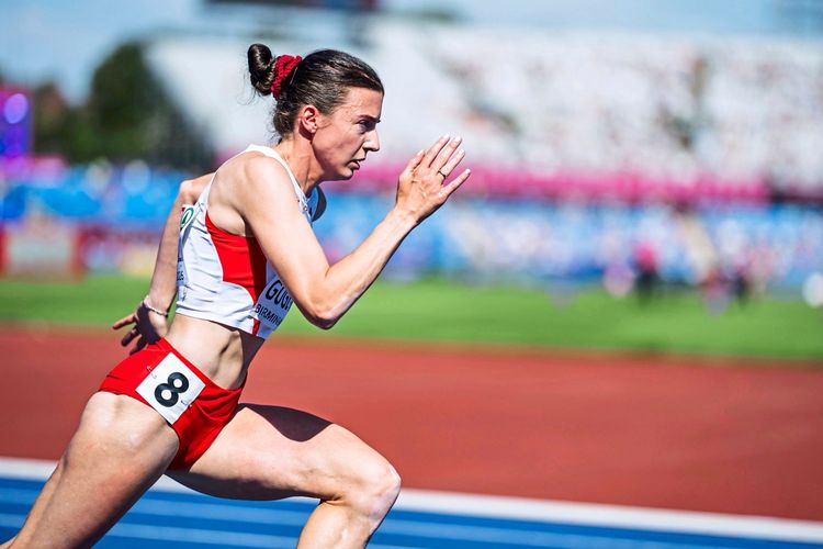 Eine Leichtathletin in Rot-Weiß sprintet während eines Wettkampfes auf einer Laufbahn. Im Hintergrund sind ein unscharfes Stadion und Zuschauer zu sehen.
