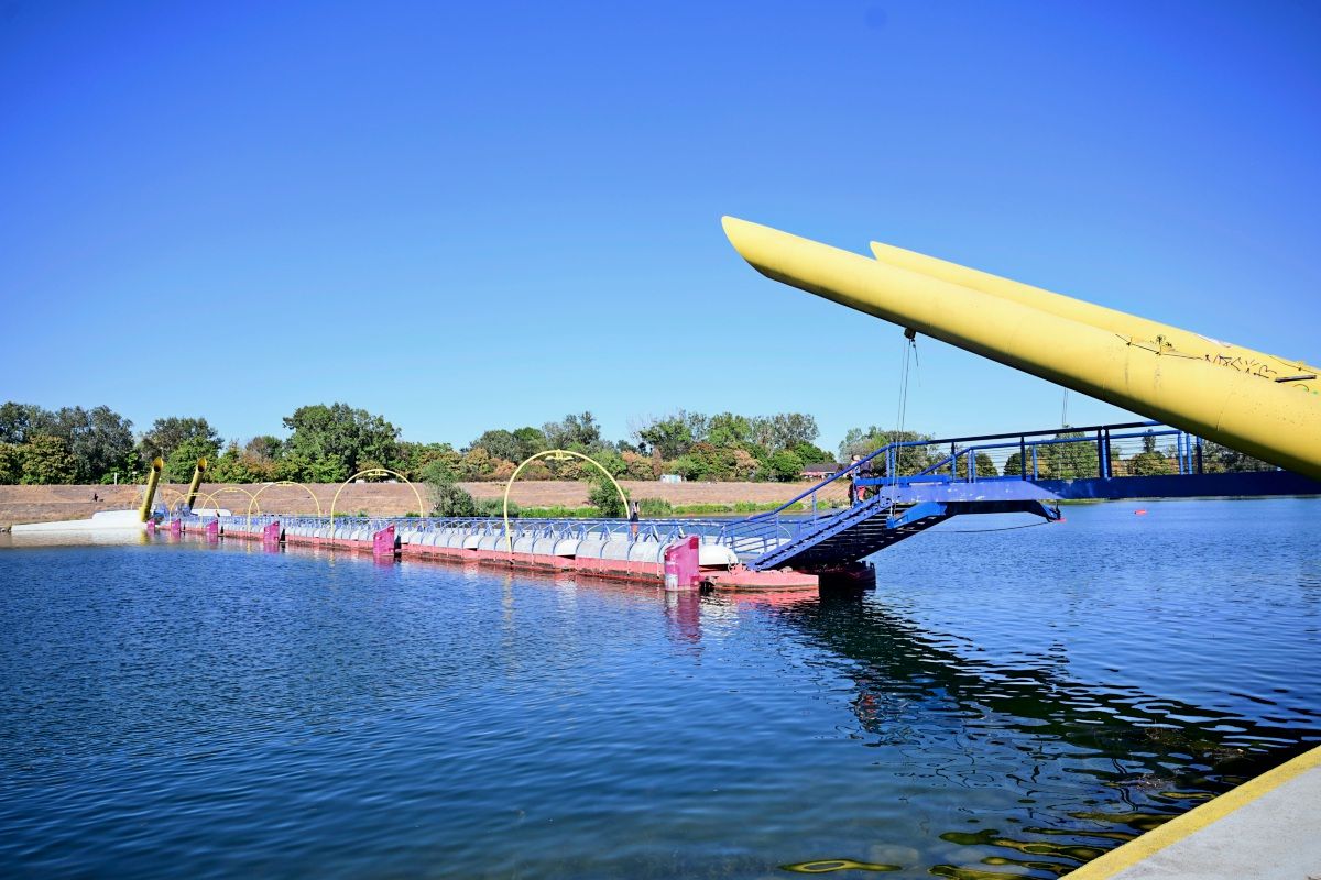 Eine bunte schwimmende Brücke mit gelben Bögen und Rampen führt über ein ruhiges Gewässer. Im Hintergrund sind Bäume und eine offene Landschaft bei klarem, sonnigem Wetter zu sehen.
