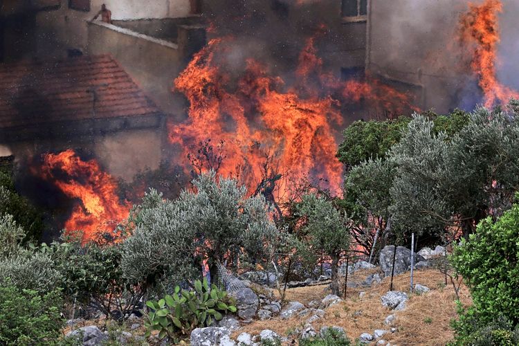 Ein intensives Feuer in einer bewachsenen Hügellandschaft nahe Wohnhäusern. Die Flammen lodern hoch und bedrohen die umliegenden Gebäude und Vegetation. Dichte Rauchwolken steigen in den Himmel.
