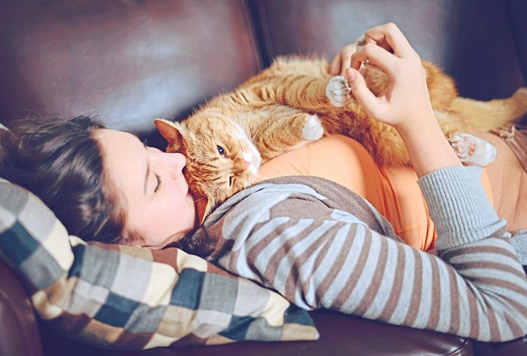 Eine Frau liegt mit einer auf ihr zusammengerollt liegenden Katze kuschelnd auf einer Couch.