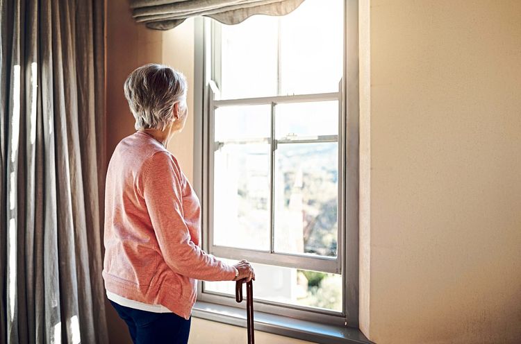 Ältere Frau mit Stock steht alleine vor Fenster und schaut hinaus