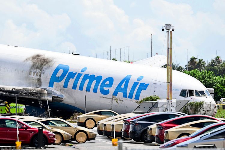Eine Prime Air-Frachtmaschine liegt beschädigt am Boden mit sichtbaren Brandspuren, nachdem sie bei der Landung am Miami International Airport am 6. September 2026 die Landebahn überrannt und Fahrzeuge getroffen hatte. Im Vordergrund stehen Tesla-Fahrzeuge auf einem Parkplatz. Im Hintergrund sind Bäume und ein klarer Himmel zu sehen.