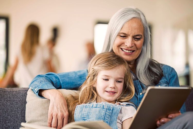 Eine ältere Frau sitzt mit einem kleinen Mädchen auf einer Couch, beide schauen gemeinsam ein Tablet an