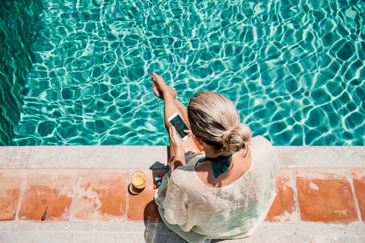 Eine Frau sitzt am Rand eines Pools, ihre Füße im Wasser. Sie hält ein Smartphone in der Hand. Neben ihr steht ein Glas mit einem Getränk.