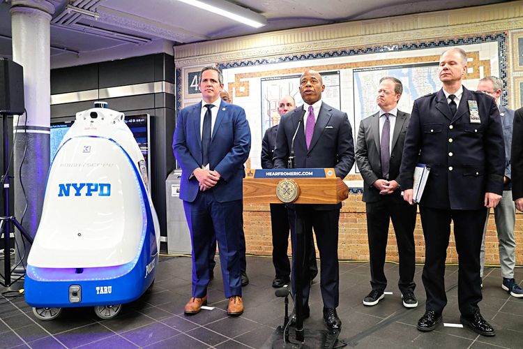 Der beinahe menschgroße Roboter ist neben dem Bürgermeister und einigen Beamten in der U-Bahn Station Times Square positioniert. Es wird eine Pressekonferenz abgehalten.
