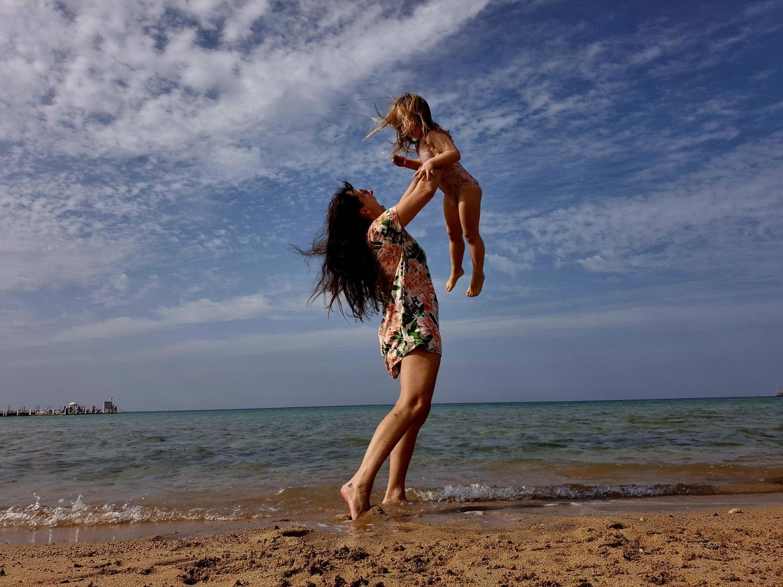 Eine Frau hebt ein Mädchen in die Luft. Die beiden Personen befinden sich am Meer an einem Sandstrand.