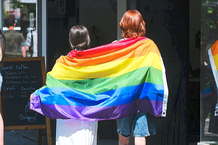 Zwei Personen stehen mit dem Rücken zur Kamera, sie sind mit einer bunten Regenbogenfahne umhüllt.