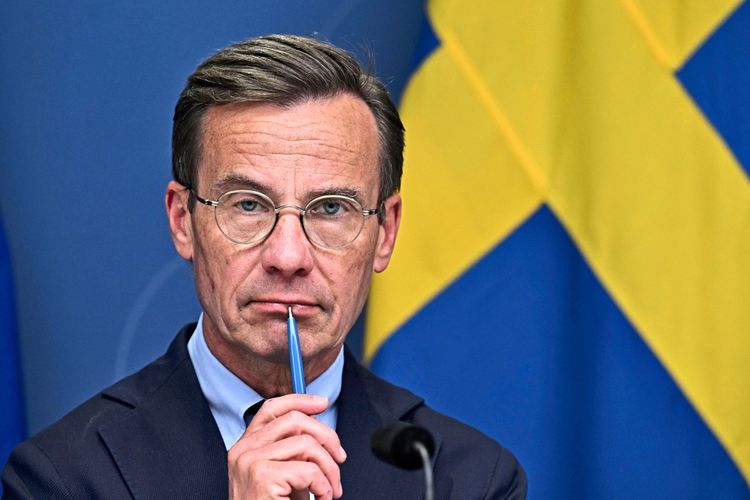 Mann in Anzug und mit Brille legt einen Kugelschreiber an seine Lippen, im Hintergrund die schwedische Flagge.