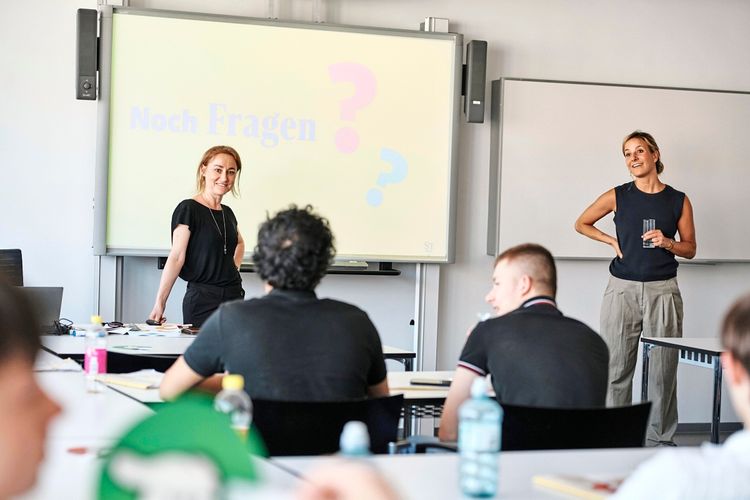 Zwei Frauen stehen vor einer Projektionsfläche in einem Seminarraum und sprechen zu einer Gruppe von Teilnehmenden. Auf der Leinwand steht „Noch Fragen?“ mit Fragezeichen. Die Teilnehmenden sitzen an Tischen und sehen zu.