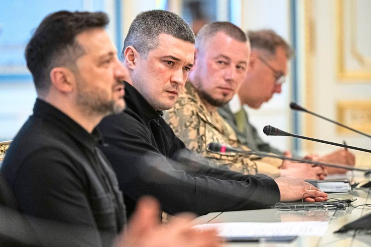 Vier Männer sitzen an einem Konferenztisch mit Mikrofonen und Unterlagen. Die Szene zeigt ein offizielles Treffen in einem prunkvoll eingerichteten Raum.