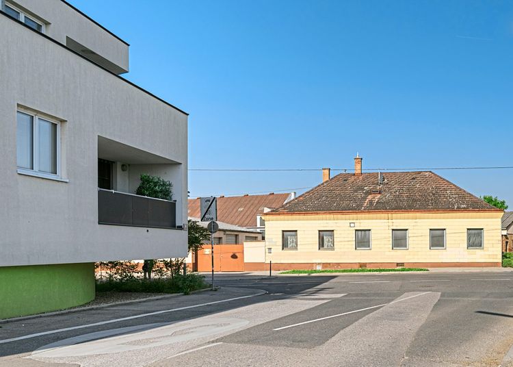 Ein modernes, geradliniges Mehrfamilienhaus steht auf einer Kreuzung mit einem alten, niedrigen Einfamilienhaus.