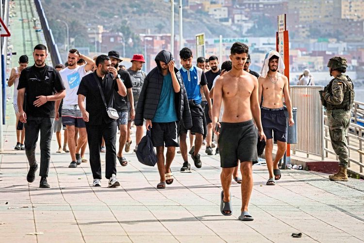 Eine Gruppe von Migranten überquert am frühen Morgen die Tarajal-Grenze von Ceuta (Spanien) zurück nach Marokko. Im Hintergrund sind Gebäude und eine person in militärischer Uniform zu sehen.