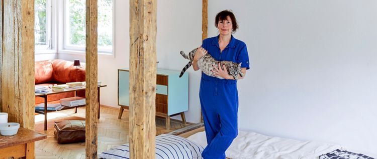 Eine Frau in einem blauen Overall steht in einem modern eingerichteten Zimmer und hält eine getigerte Katze im Arm. Im Hintergrund sind ein Sofa, ein Tisch mit Büchern und ein türkisfarbener Schrank zu sehen. Holzbalken trennen den Schlaf- und Wohnbereich.