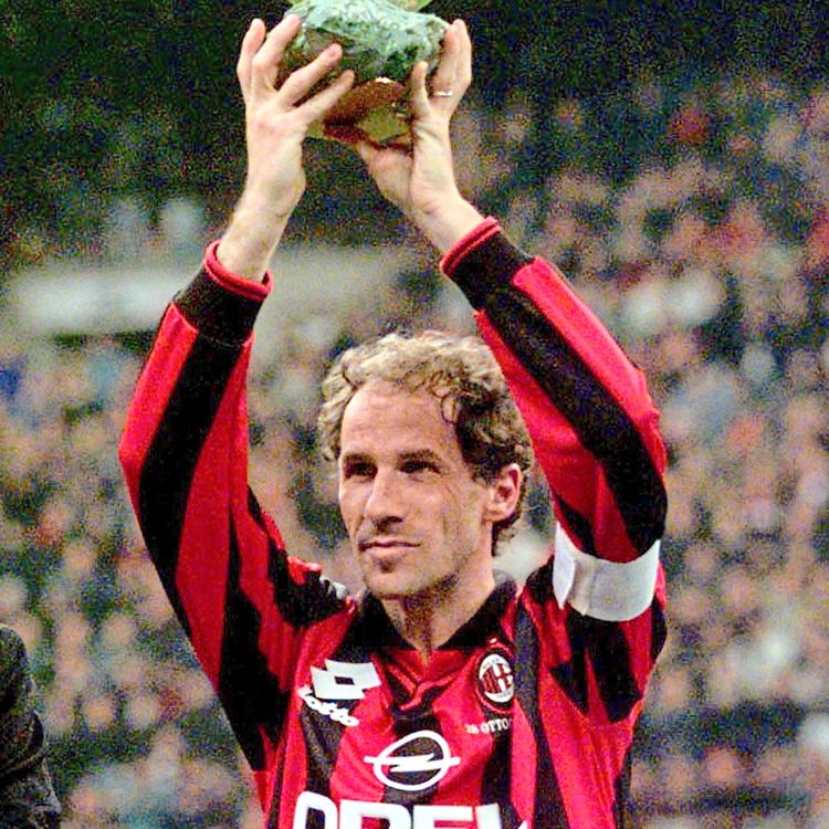 Ein Fußballspieler im rot-schwarz gestreiften Trikot von A.C. Milan hält einen goldenen Ball-Trophäe über seinem Kopf in einem Stadion.