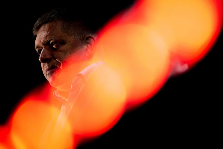 Robert Fico bei einer Wahlkampfveranstaltung. Der Politiker ist in der linken Bildhälfte zu sehen, vor schwarzem Hintergrund spiegelt sich orange-rötliches Licht.