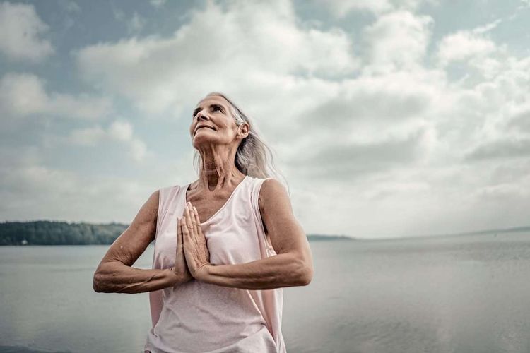 Ältere Frau mit langen weißen Haaren meditiert vor einem See