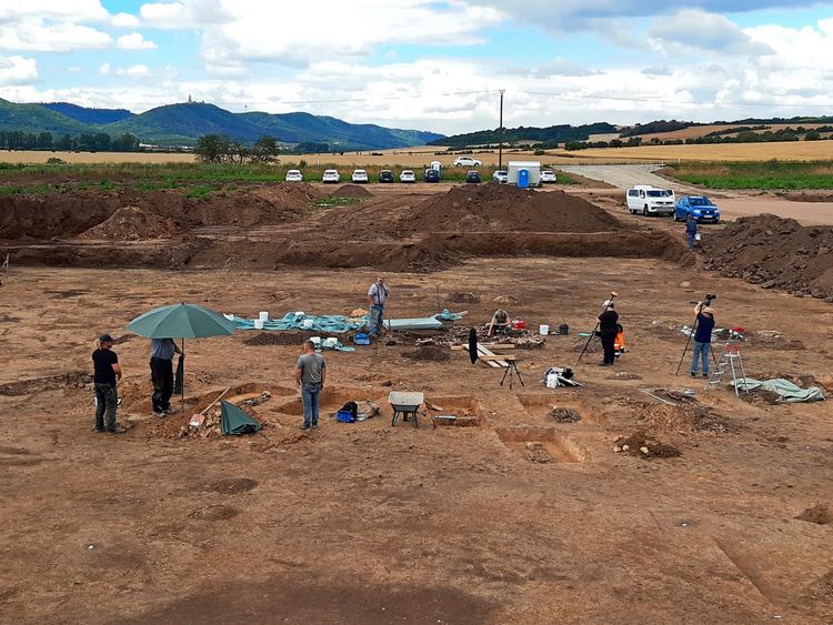 Archäologische Ausgrabung eines Gräberfeldes bei Brücken in Ostdeutschland. Mehrere Personen arbeiten an freigelegten Gräbern auf einer braunen Erdfläche unter freiem Himmel. Im Hintergrund sind Autos, Hügel und Felder zu sehen.