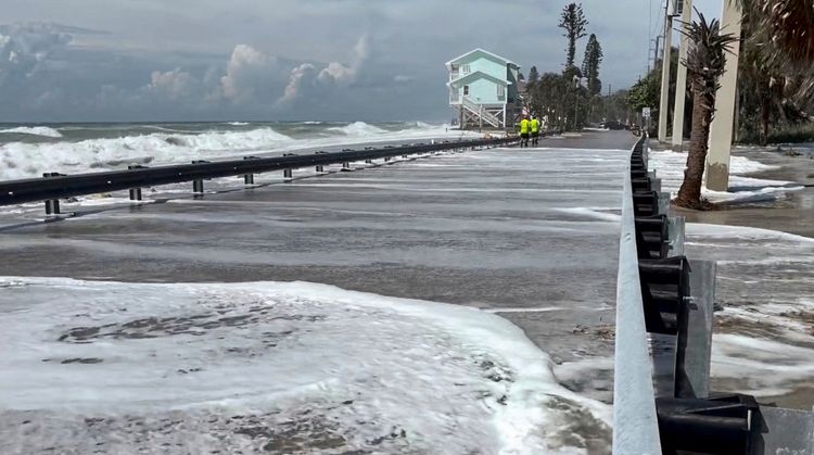 Menschen gehen entlang einer Küstenstraße in Englewood, Florida, während Wellen die Straße überfluten. Im Hintergrund ist ein an der Küste stehendes Haus zu sehen. Der Himmel ist bewölkt, und Sturmbedingungen deuten auf die bevorstehende Ankunft von Hurrikan Isaias hin.