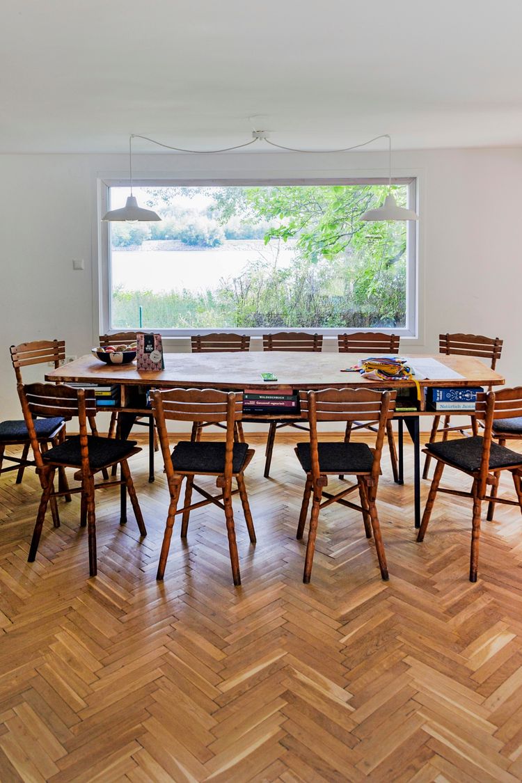 Ein Esszimmer mit einem großen Holztisch, umgeben von acht Holzstühlen mit dunkler Polsterung. Auf dem Tisch liegen Bücher, Dekorationsgegenstände und ein Spielzeug. Im Hintergrund bietet ein großes Fenster einen Blick auf eine naturnahe Landschaft mit Wasser und Bäumen. Der Boden ist mit einem Fischgrät-Holzparkett ausgelegt.