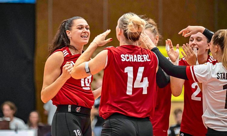 Eine Gruppe von Volleyballspielerinnen in roten und weißen Trikots feiert gemeinsam auf dem Spielfeld. Eine Spielerin mit der Nummer 18 lächelt, während sie ihre Teamkolleginnen abklatscht. Im Hintergrund sind unscharf Zuschauer und der Hallenbereich zu erkennen.